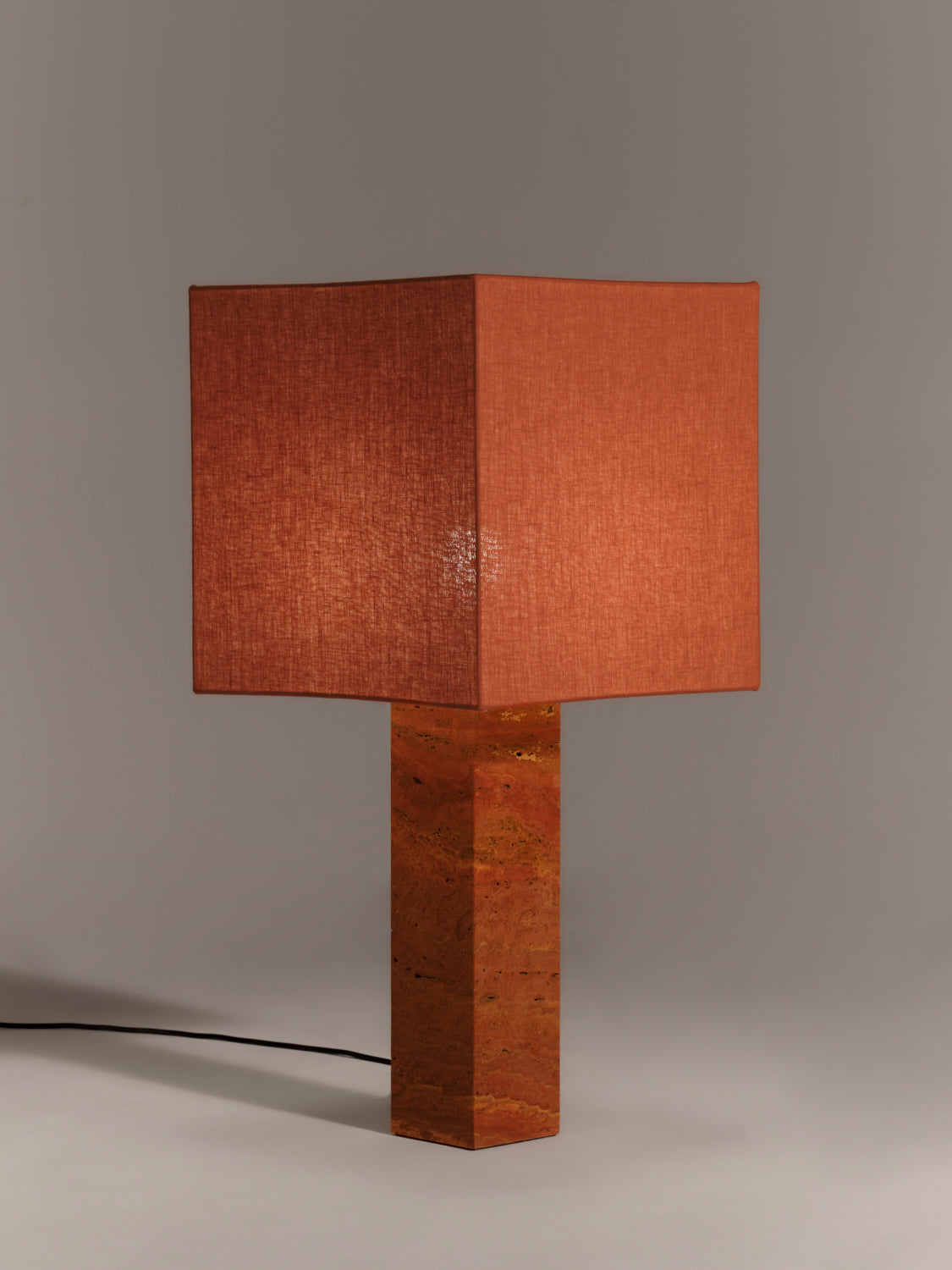 Table lamp with a square rust red linen shade and square red travertine base on a gray background