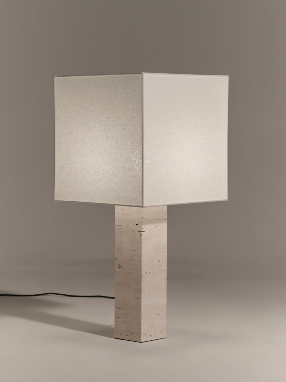 Table lamp with a square white linen shade and square white travertine base on a gray background