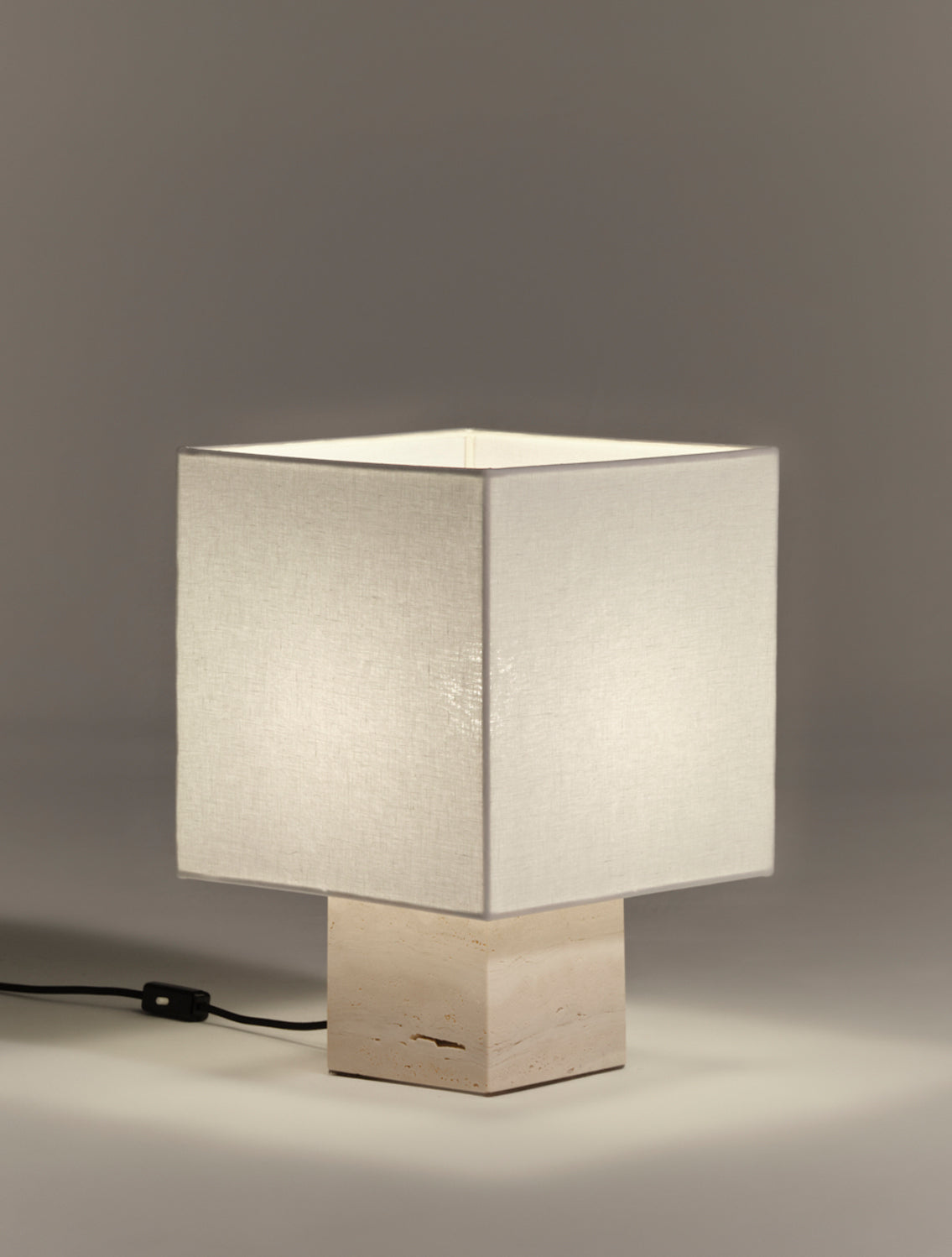 Table lamp with a square white linen shade and square white travertine base on a gray background