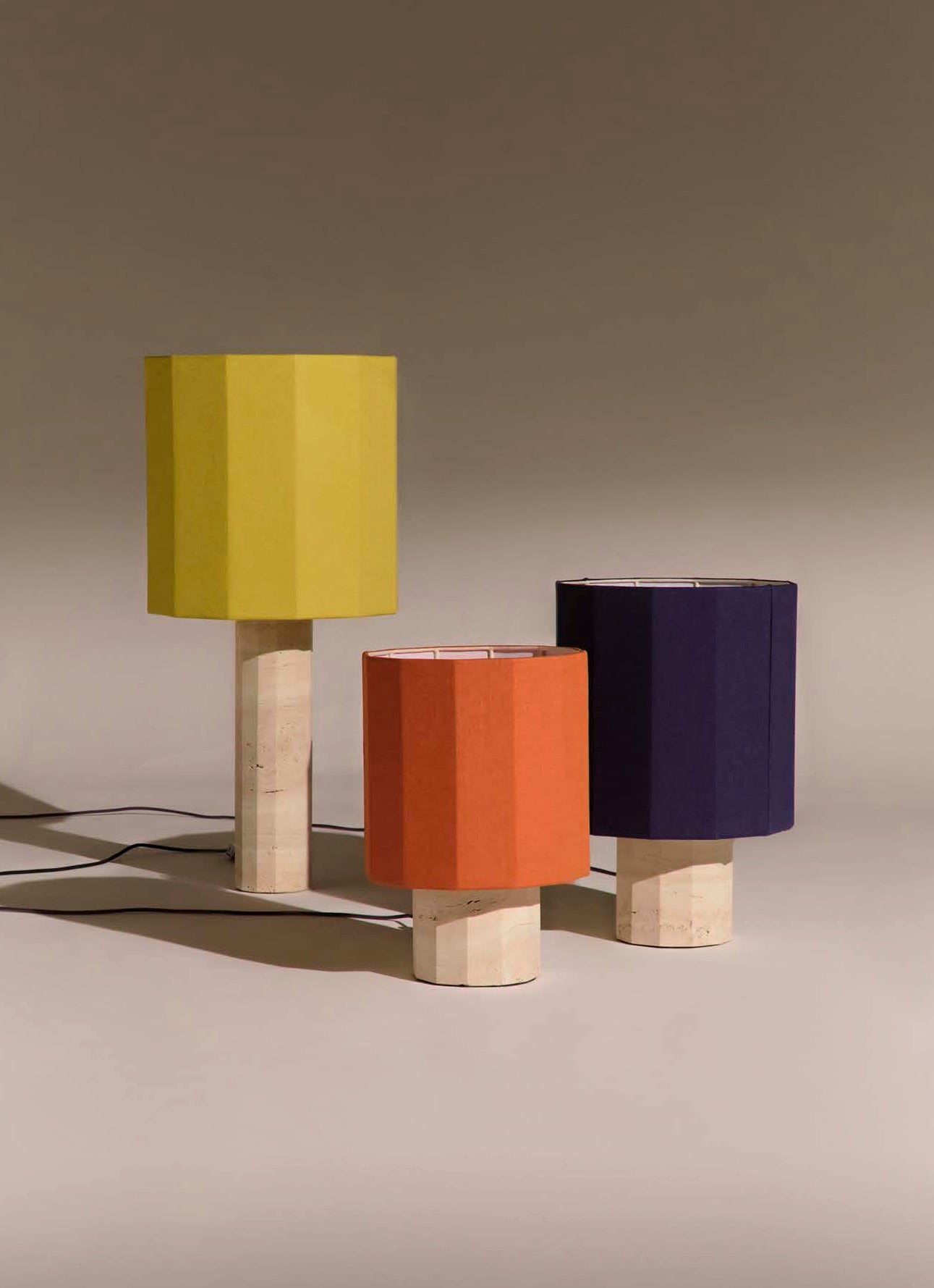 Three white travertine fluted dodecagon table lamps on a neutral background with shades in yellow, rust, and navy blue
