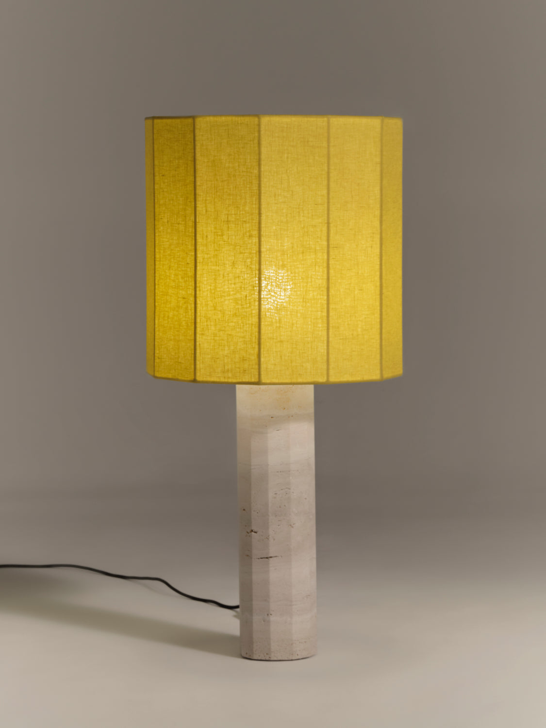 Table lamp with a fluted dodecagon yellow linen shade and matching white travertine base on a gray background