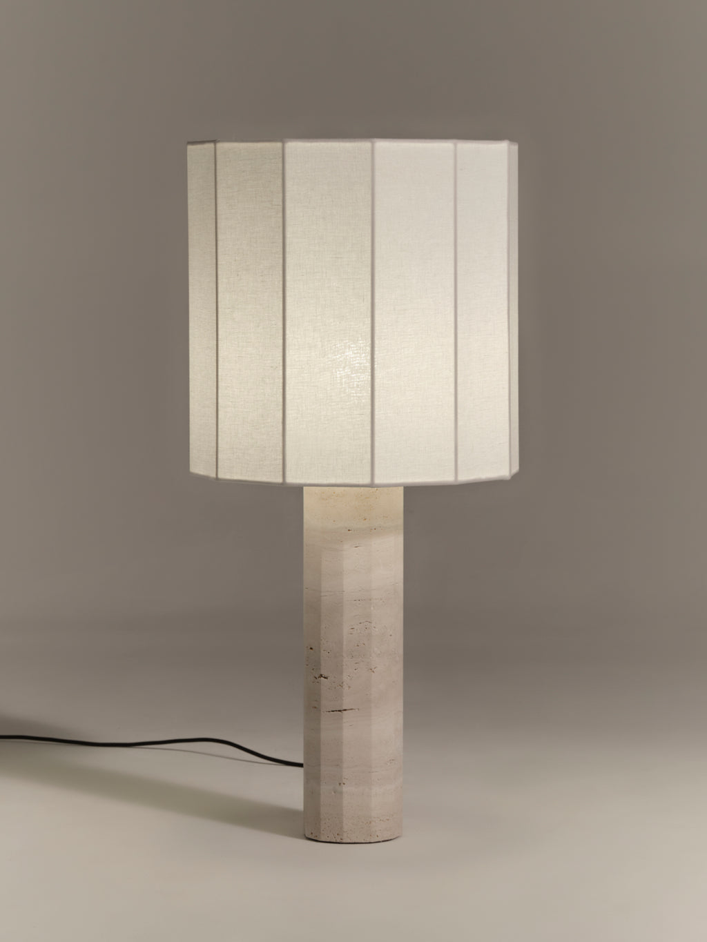 Table lamp with a fluted dodecagon white linen shade and matching white travertine base on a gray background