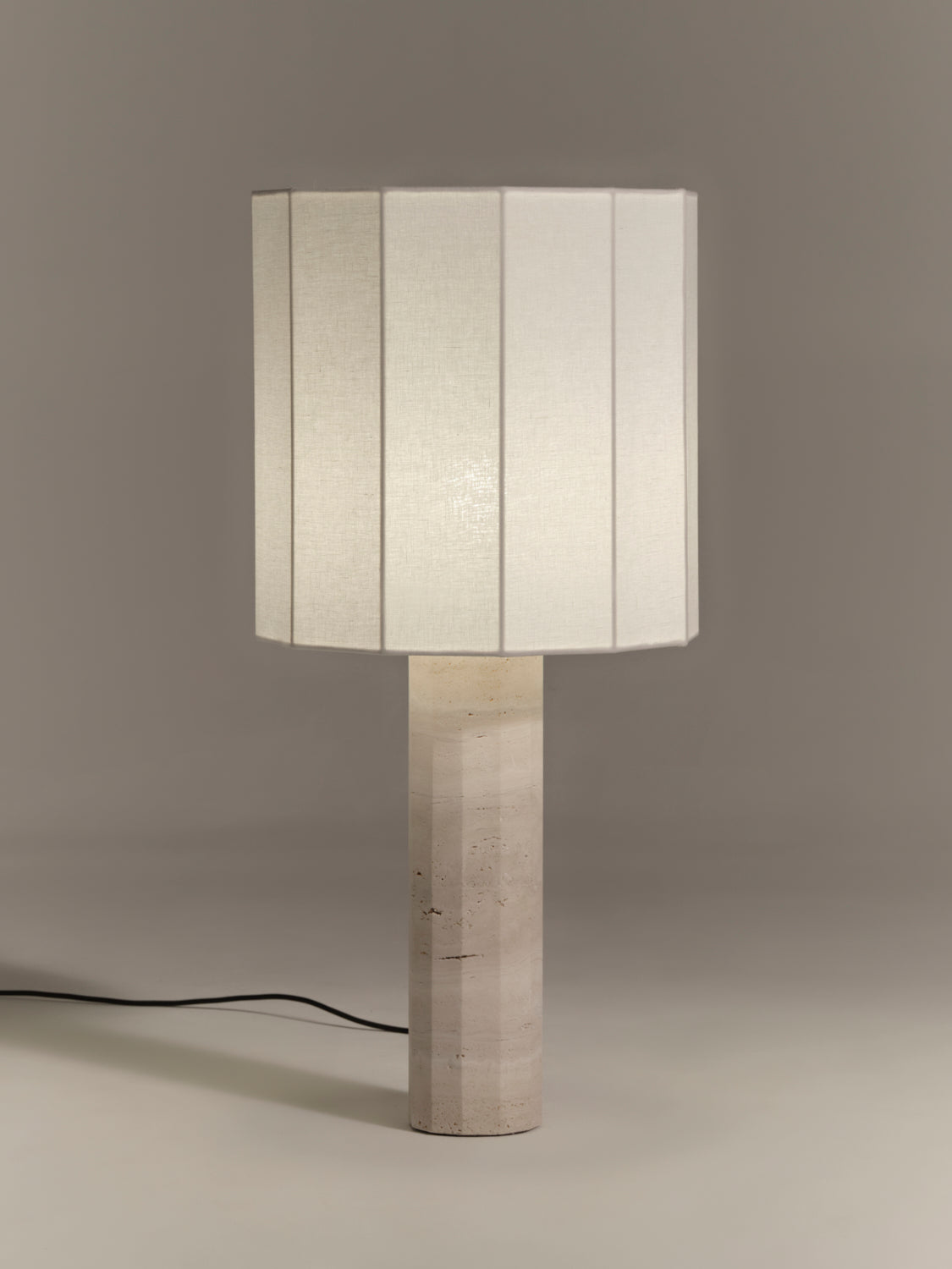 Table lamp with a fluted dodecagon white linen shade and matching white travertine base on a gray background