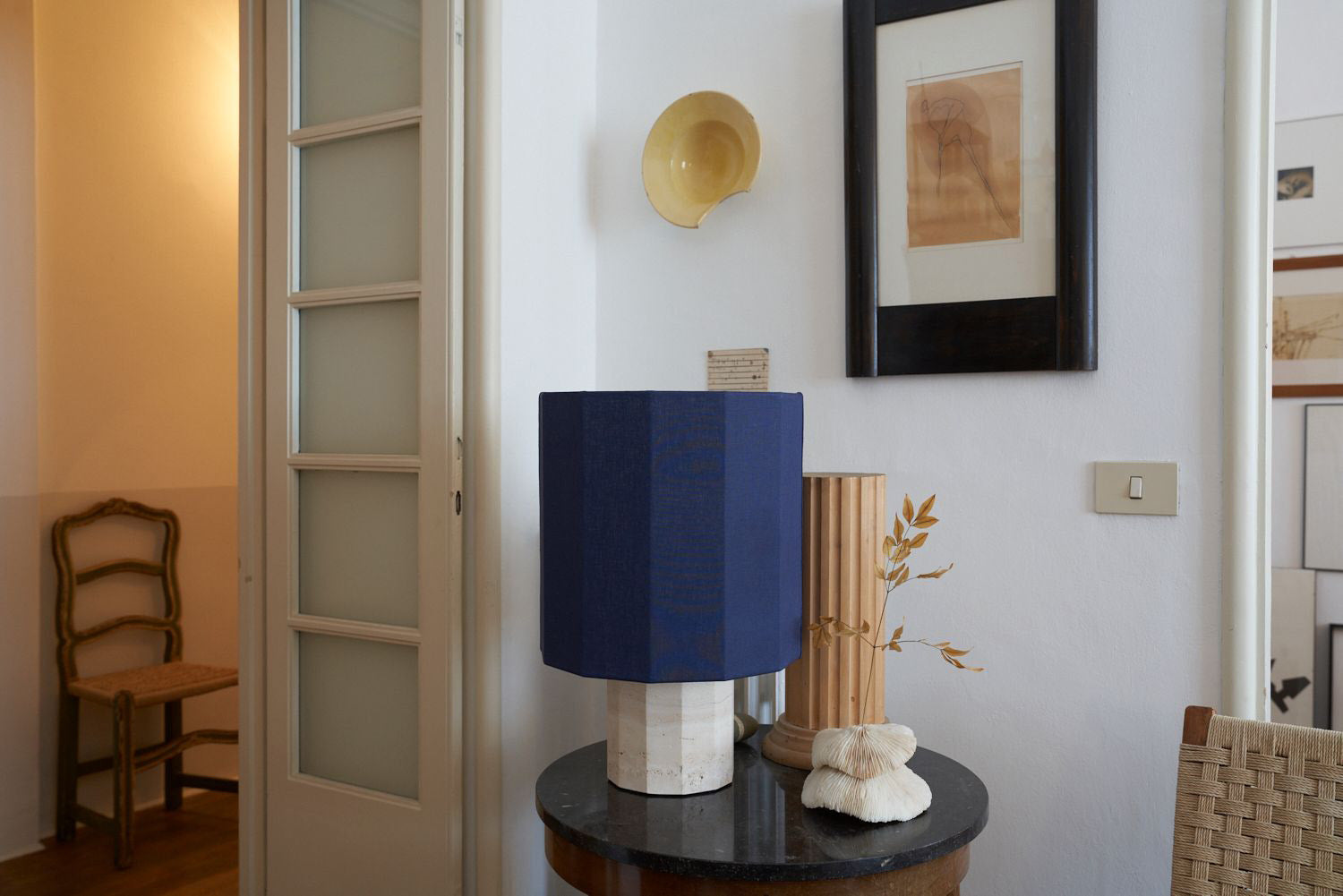 Navy blue lamp on a round table with a chair and wall art in the background