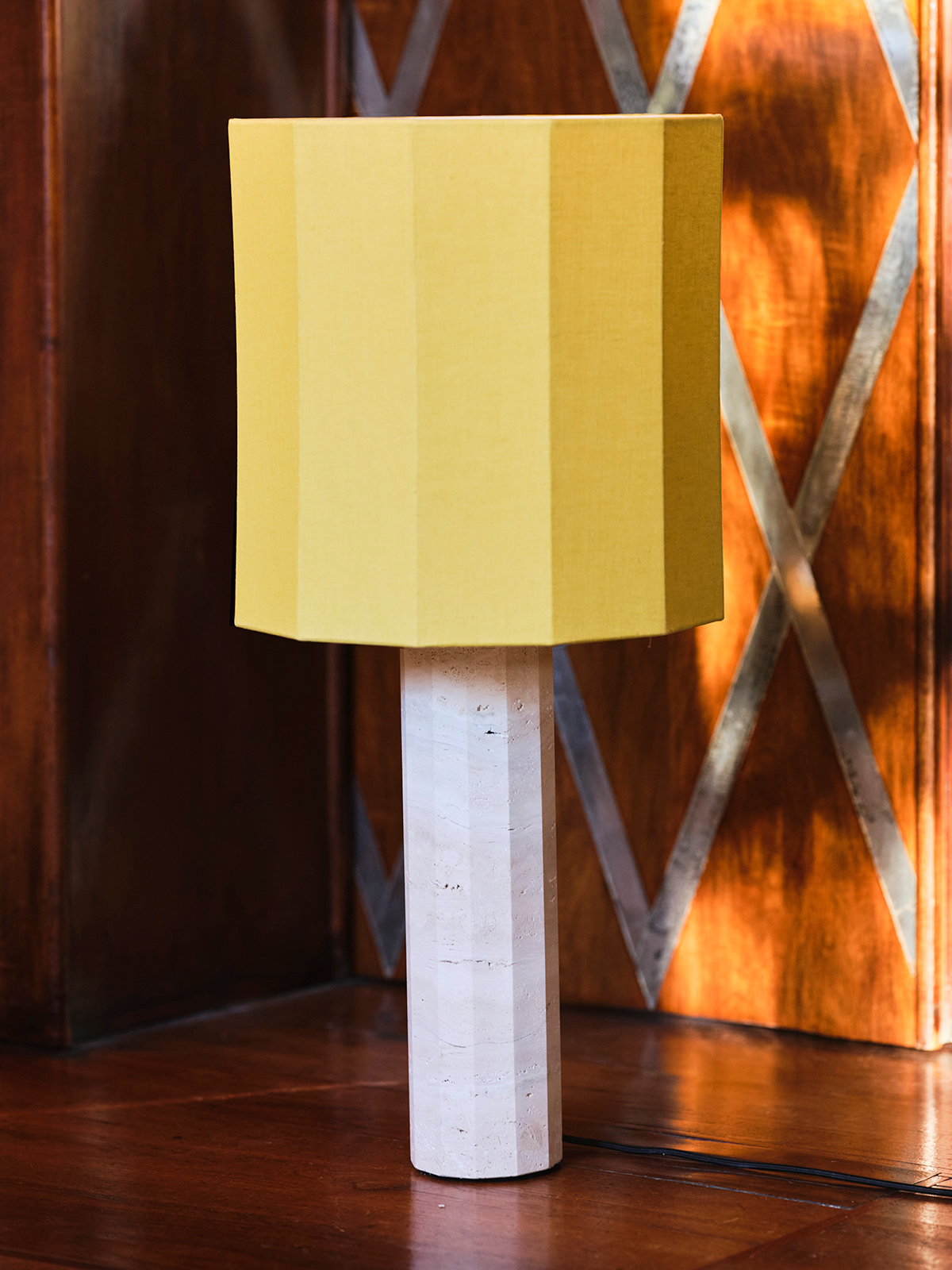 Yellow lampshade on a white travertine fluted base against a decorative wall.