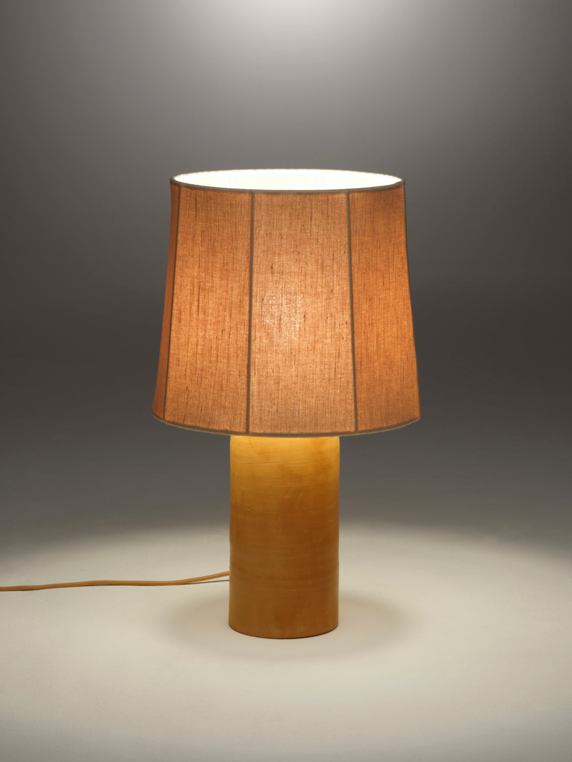 Lit table lamp with a fluted caramel linen shade on a terracotta rounded base against a gray background