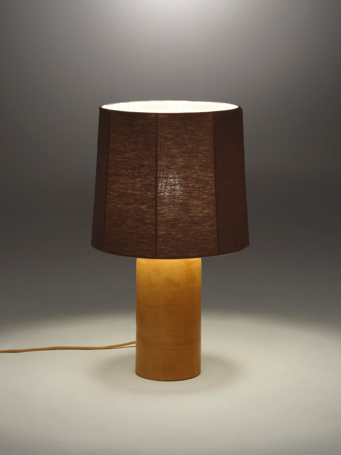 Lit table lamp with a fluted chocolate linen shade on a terracotta rounded base against a gray background