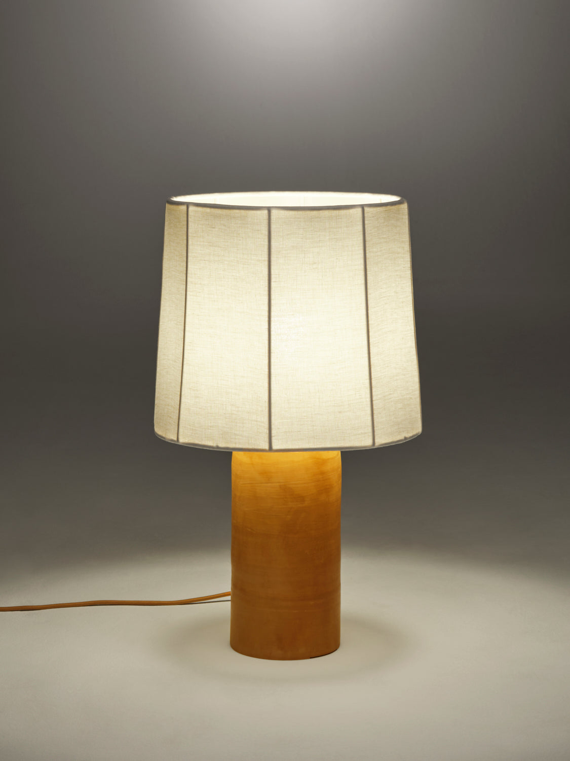 Lit table lamp with a fluted white linen shade on a terracotta rounded base against a gray background